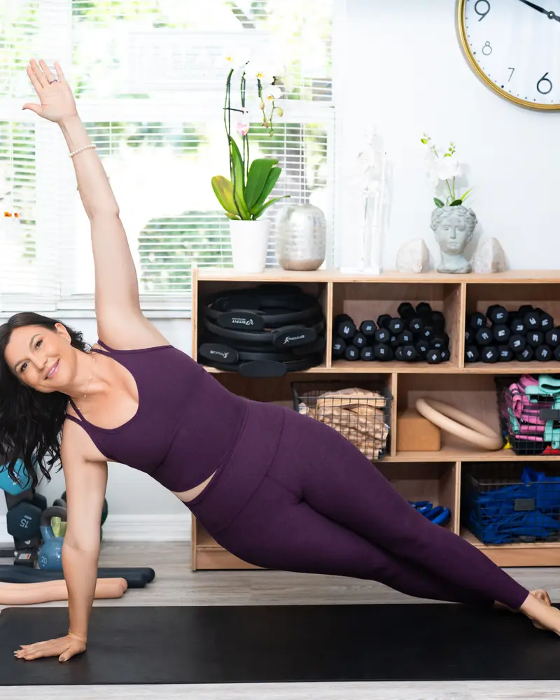 Leia holding a side plank on a mat in the Pilates Port Charlotte studio, weights and props on shelves behind her